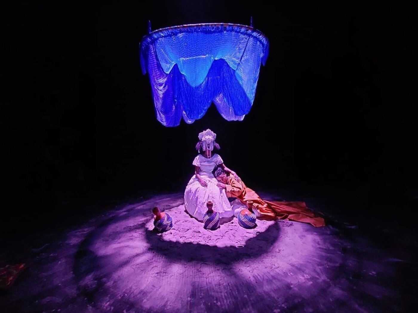 Bühnenaufnahme in dunkler Umgebung mit starkem violett-blauem Licht. In der Mitte sitzen zwei Performerinnen auf einer hellen, sandartigen Fläche. Eine Person trägt ein weißes, historisch anmutendes Kostüm mit Kopfschmuck und sitzt aufrecht; eine zweite Person liegt seitlich an sie gelehnt und ist in orangefarbenen Stoff gehüllt. Über ihnen hängt ein großes, blau beleuchtetes, textilartiges Objekt, das wie ein schwebender Baldachin wirkt. Vor den Performerinnen stehen mehrere runde Gefäße. Der Bühnenraum ist sonst fast vollständig schwarz und lenkt den Blick auf die zentrale Szene.