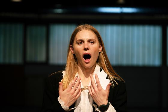 A woman in a theatrical costume stands in a large hall, her mouth wide open in an exaggerated expression, as if she is about to speak or shout.