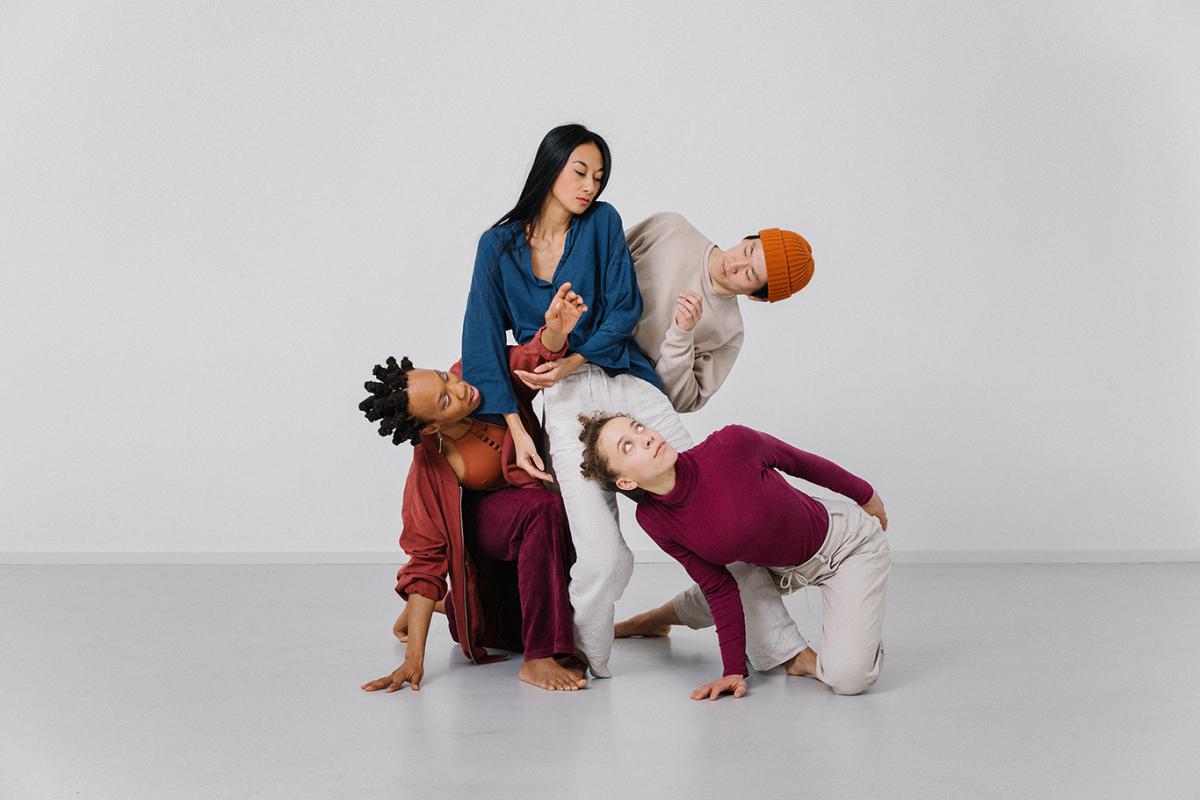 Four performers are arranged in a grouped, sculptural pose. Three are supported close to the floor while one sits above them. Their bodies form an interwoven, dynamic composition against a neutral background.