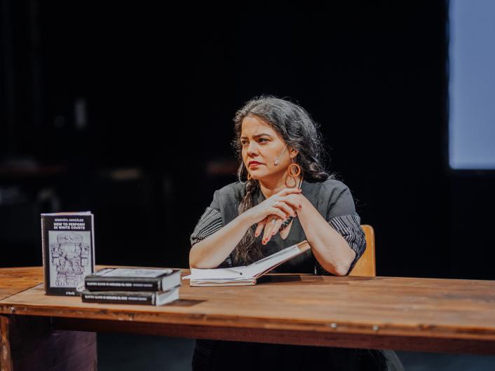A person sits at a wooden table with arms crossed, looking thoughtfully to the side. Books and notes lie on the table, the stage behind them is dark.