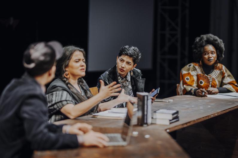Several people sit at a long table engaged in discussion. One person gestures with their hands while others listen attentively. Books, laptops, and notes are spread across the table.