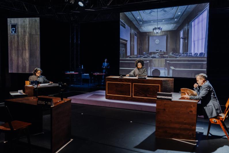 A stage scene depicts a recreated courtroom with several people at wooden desks. A large screen in the background shows an image of a courtroom.