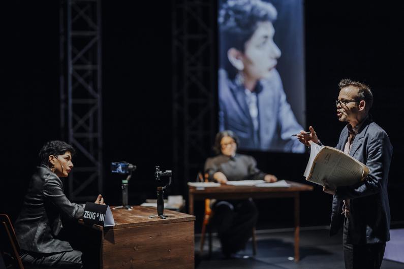 Eine Person steht mit Notizen in der Hand und spricht zu zwei anderen, die an Tischen sitzen. Im Hintergrund ist ein vergrößertes Porträt einer der sprechenden Personen auf einer Leinwand zu sehen.