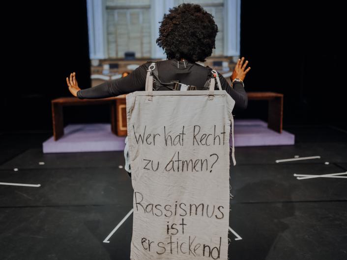 A person stands with their back to the camera on stage. A fabric banner on their back reads: “Who has the right to breathe? Racism is suffocating.”