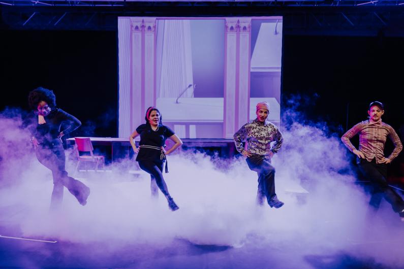 Vier Personen stehen in einer Nebelwolke auf der Bühne und führen eine Tanzszene auf. Der Hintergrund ist in violettes Licht getaucht.