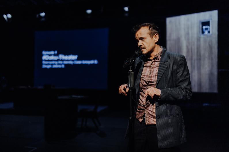 A person stands at a microphone speaking on stage. Behind them, a screen displays white text on a dark background.