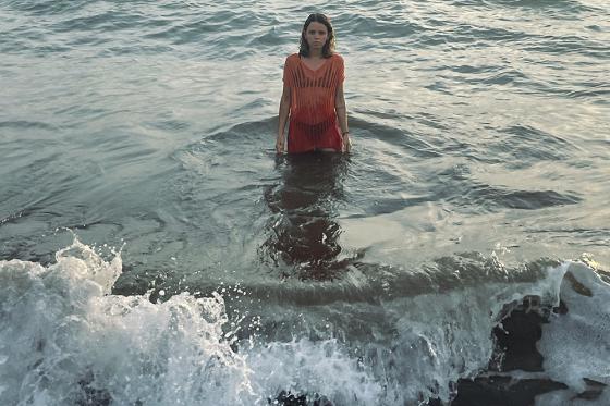 "Eine Frau steht im Meer und trägt ein rotes Kleid, das Wasser reicht ihr bis zu den Oberschenkeln. Im Vordergrund brechen sanft die Wellen, und am Horizont ist ein wolkiger Himmel zu sehen."