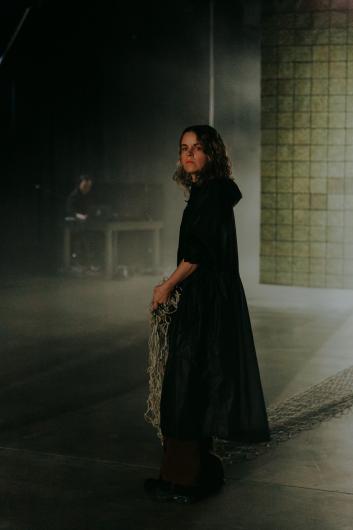 A woman in a black coat stands in a foggy, dimly lit setting. She holds a net in her hand, while a person is seated at a table in the background.