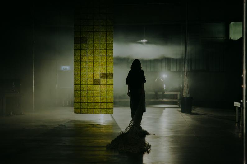 A silhouette of a person in a long black dress stands in a dark room. In front of them hangs a large greenish shimmering grid, and another person is seated in the light in the background.