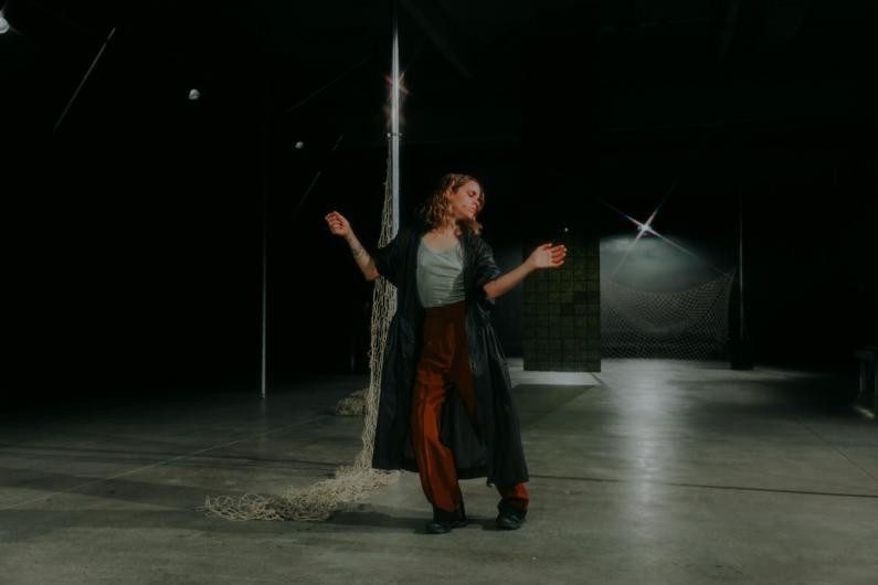 A woman with wavy brown hair wearing a long black coat, brown pants, and a light green top poses with arms outstretched in a dimly lit industrial space with netting and metallic poles