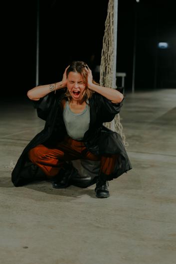 A woman crouches with her hands on her head, screaming with her mouth wide open, in an industrial space with netting visible in the background.
