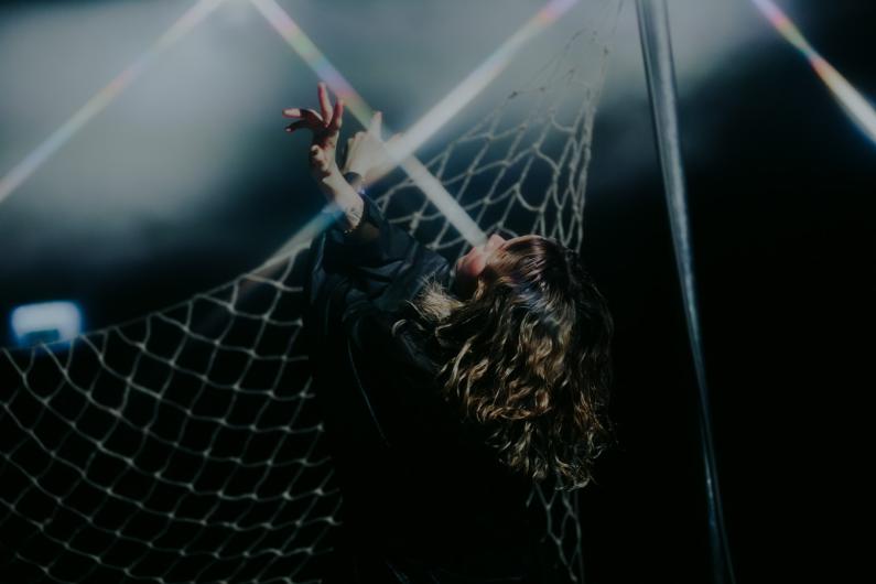 A woman with wavy hair reaches upward while leaning against a net under dramatic lighting, with rainbow flares crossing the frame.