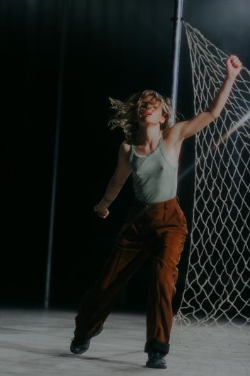 A woman with loose, wavy hair is performing energetically in front of a net. She wears a light green tank top and brown pants, raising one arm upwards while caught in dynamic motion. The stage lighting emphasizes her intensity.