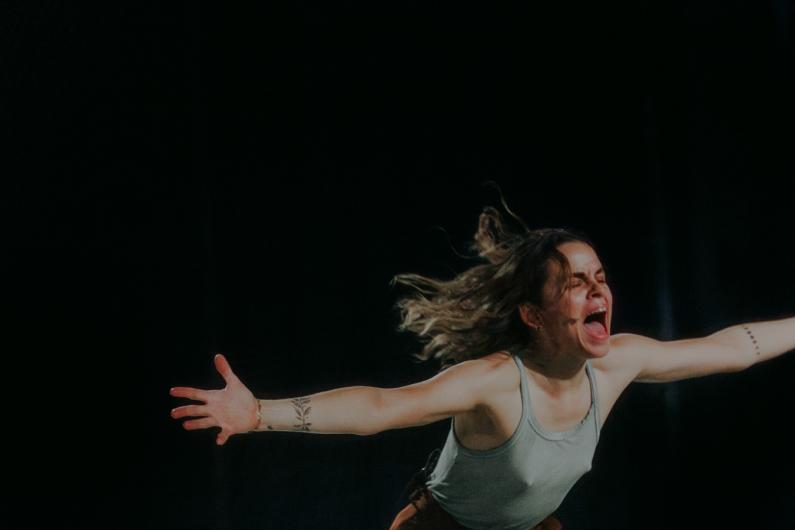 A woman, her face expressing raw emotion, extends both arms wide while shouting. Her hair flows backward as she moves on a dark stage, wearing a light green tank top and brown pants.