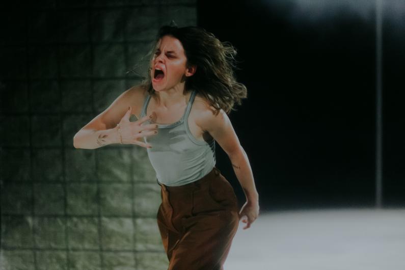 A woman clutches her chest with one hand, her mouth open in a scream, expressing intense emotion. She wears a light green tank top and brown pants, standing in front of a textured backdrop on a dimly lit stage.