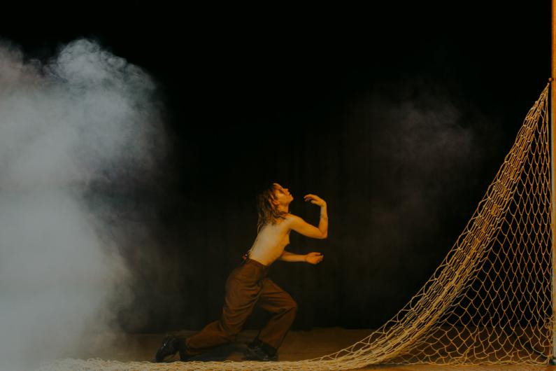 A person kneels on the ground in a dramatically lit scene. They are topless, wearing brown trousers, and pose with a net against a dark background.