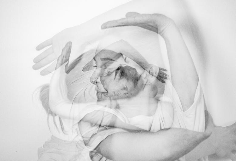 Eine schwarz-weiß Foto-Collage zeigt den Oberkörper einer Frau. Die Frau hat weiße Haut, trägt ein weißes T-Shirt und hat ihr Haare zu einem Zopf gebunden. Sie bildet mit ihren Armen und Händen Linien, Umrahmungen und Formen vor und um ihr Gesicht. Es sind in diesem Bild drei Fotos unterschiedlicher Positionen übereinandergelegt, was einen verschwimmenden Effekt erzeugt. Es sind viele Arme, Gesichter und Hände erkennbar, und doch gehören sie alle zusammen.