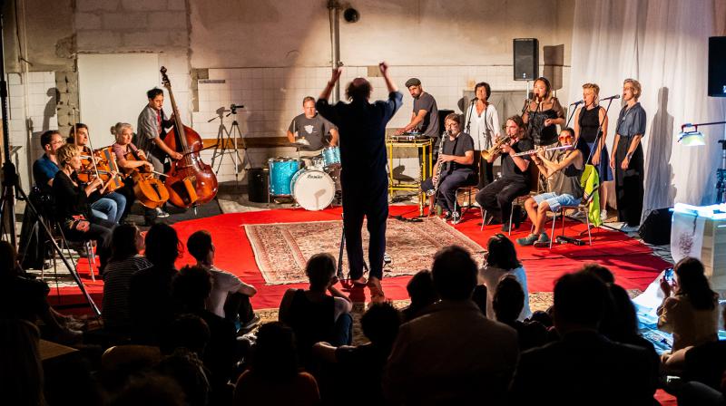 Indoor musical performance with a conductor leading an ensemble of string, percussion, brass, and woodwind musicians and vocalists on a red carpet. Audience members are seated on the floor, watching intently. The room has unfinished walls, adding an industrial aesthetic.