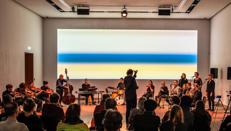 Concert performance with a conductor leading an ensemble of musicians and vocalists in a modern gallery space. The ensemble includes string, keyboard, percussion, brass, and woodwind players. A large abstract artwork with gradient colors in blue, white, and yellow fills the background wall. Seated audience members observe the performance.