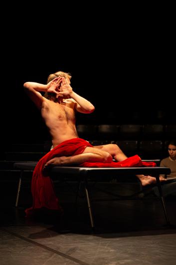 A person sits naked on a table, partially draped with red fabric. They hold their hands in front of their face, forming a triangle. The background is dark, with another person faintly visible in the distance.