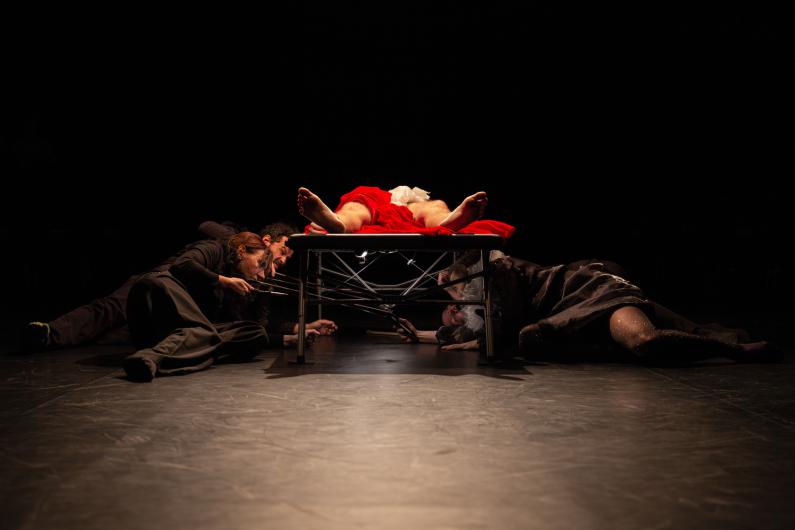 A person lies on a table, covered with red fabric. Beneath the table, several individuals in dark clothing crawl, appearing to hold tools or rods. The scene is dramatically lit and set in a dark room.