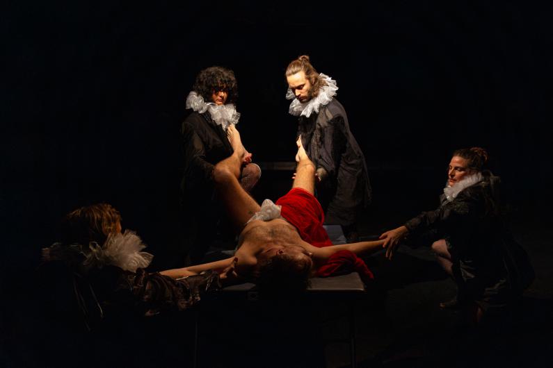 Four individuals in baroque-style clothing with white ruffled collars surround a table where a naked person lies, partially covered with red fabric. The scene is staged in a dark room with dramatic lighting on the figures.