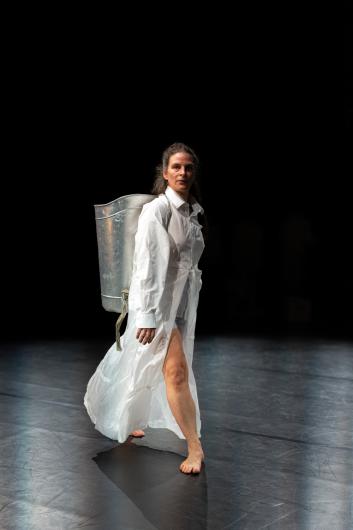 A barefoot performer in a translucent white robe and shirt carries a metallic container on their back, standing on a dimly lit stage.