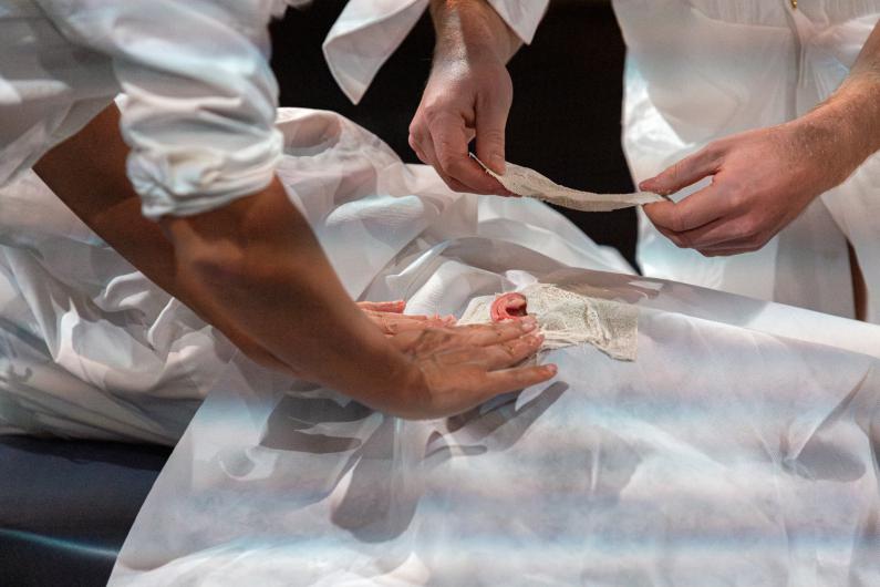 Close-up of two hands applying a bandage to an object wrapped in translucent white fabric on a dark background.