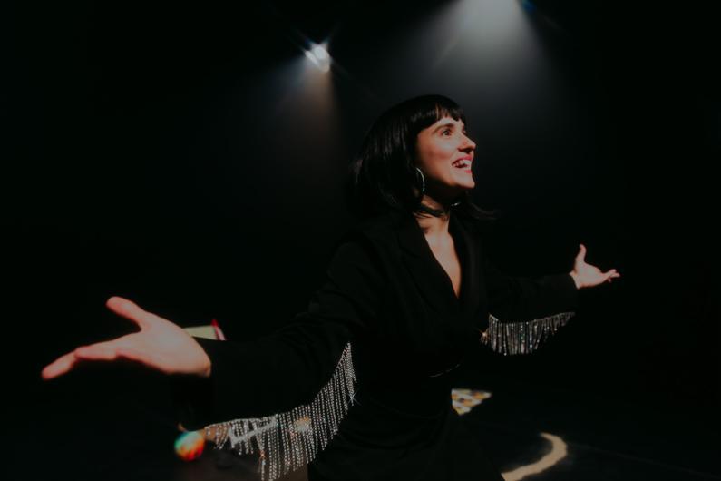 A performer in a dark setting, illuminated by spotlights, stands with open arms, smiling enthusiastically. She wears a black outfit with sparkling fringe details on the sleeves.