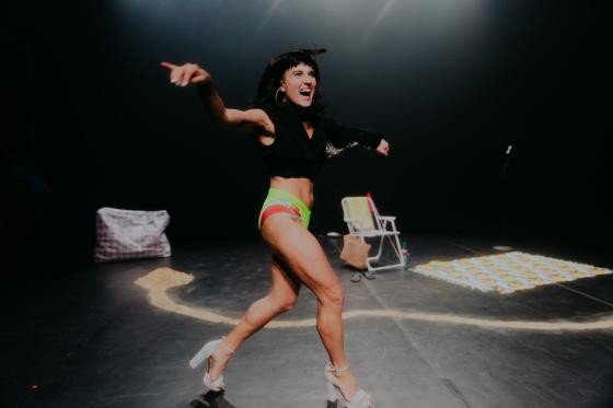 A performer in a dynamic pose on stage, appearing energetic and expressive. She is wearing neon green shorts and silver high heels, with a black top. Props such as a woven bag and a striped towel are visible in the background.