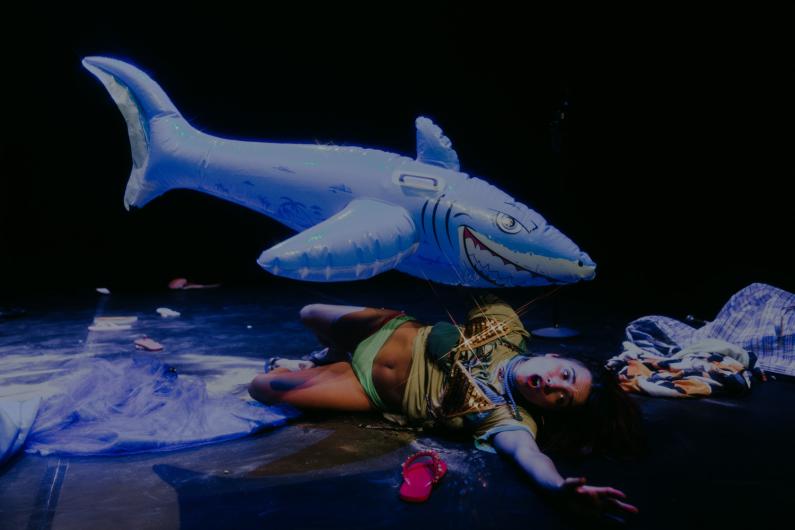 A performer lies dramatically on the floor of a dark stage, one arm outstretched, with a surprised facial expression. Above them floats a large inflatable shark with a fierce expression. The scene is set with scattered props, including a single red flip-flop, fabrics, and nets. The lighting creates a mystical, almost surreal atmosphere.