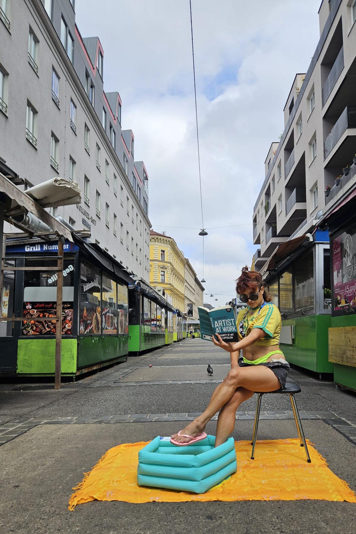Eine Frau sitzt auf einem Hocker mitten auf einer verlassenen Markstraße zwischen zwei Reihen von grün-schwarzen Marktständen. Sie trägt ein gelb-grünes Shirt, eine dunkle Shorts und Flip-Flops und hat eine Sonnenbrille auf. Sie liest ein Buch mit dem Titel "Artist at Work" und hat ihre Füße in einem kleinen, türkisfarbenen aufblasbaren Pool, der auf einer orangefarbenen Decke steht. Die Gebäude an beiden Seiten sind modern und grau, während eines im Hintergrund gelb ist. Eine Taube ist auf der Straße zu sehen, und der Himmel ist bewölkt.