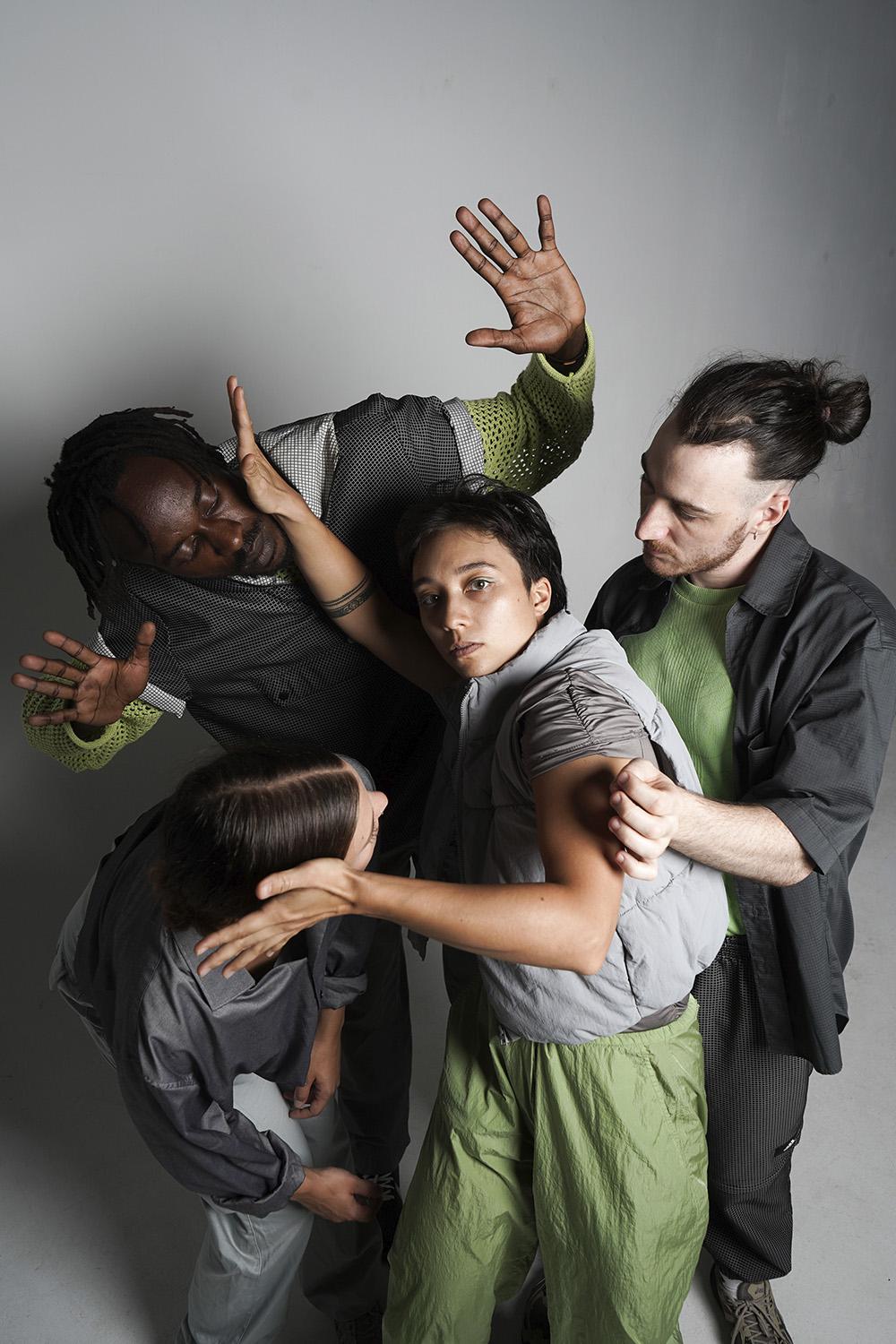 Vier Personen in dynamischen Posen, die eng miteinander interagieren. Die Szene scheint einen Moment physischer Spannung oder Ausdruck darzustellen, mit ausgestreckten Händen und Körpern, die sich aneinander lehnen. Die Personen tragen geschichtete, legere Kleidung in Grau-, Grün- und Schwarztönen.