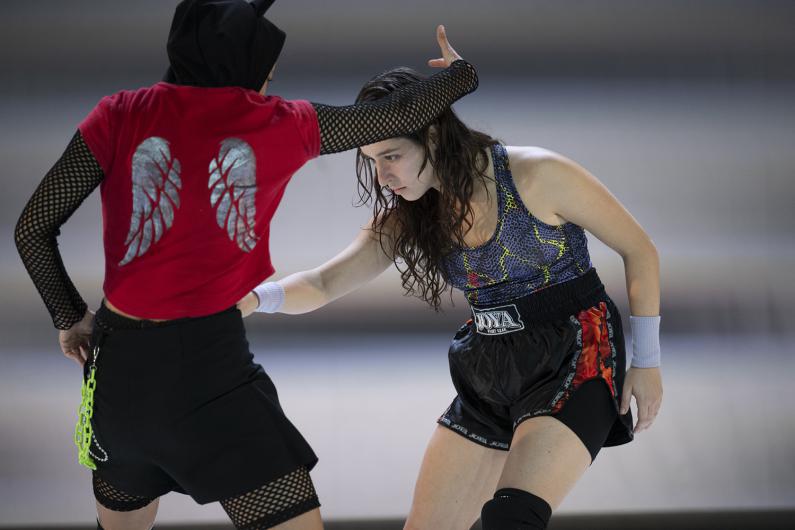Two female performers are engaged in what appears tob e an intense martial arts match. One performer, on the left, wears a red shirt with silver wing prints on the back, black mesh sleeves, and a hood. The other performer, on the right, wears a colorful athletic top and black shorts with a “Joya” logo. Both appear focused and in mid-motion against a blurred background.