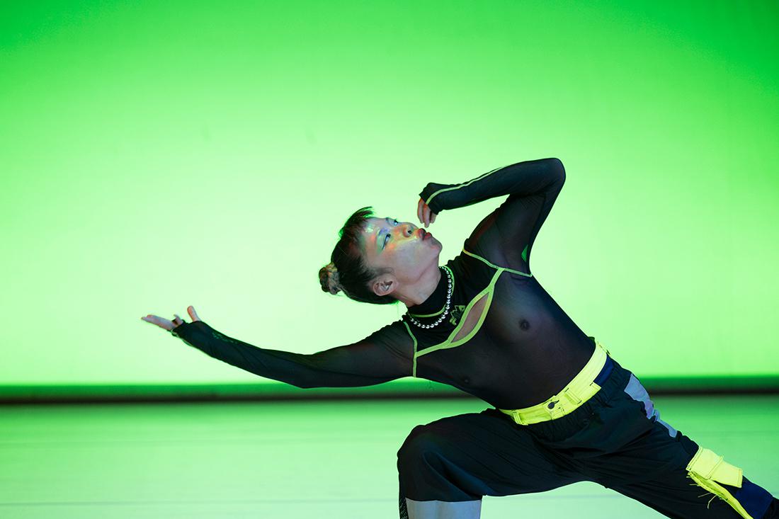 Eine Person führt eine Tanzpose vor einem leuchtend grünen Hintergrund aus. Sie trägt ein schwarzes durchsichtiges Oberteil mit neonfarbenen Akzenten und eine Hose mit neonfarbenen Details. Ihr Gesicht ist mit buntem Make-up bemalt, und sie scheint eine ausdrucksstarke Geste zu machen.