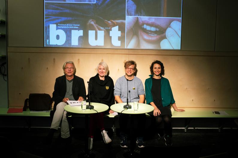 Vier Personen sitzen bei einer Presse- oder Gesprächssituation an zwei kleinen runden Tischen vor Mikrofonen und Wassergläsern. Im Hintergrund ist eine großformatige Projektion mit einem brut-Sujet zu sehen. Darauf steht unter anderem: „Find brut all over Vienna!“, „Karlsplatz 5, 1010 Wien“, „brut-wien.at“ und groß das Logo „brut“.