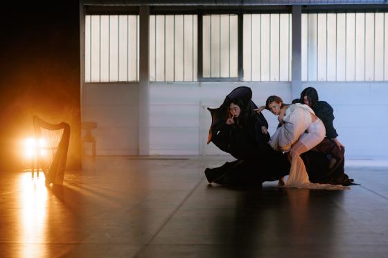 Drei Performer*innen in einer weiten, leeren Halle bilden gemeinsam eine skulpturale, bodennahe Pose. Zwei Personen tragen schwarze, fliessende Stoffe, eine Person in der Mitte trägt ein helles, weisses Outfit mit voluminösen Ärmeln. Links im Bild steht eine Harfe vor einer grellen, warmen Lichtquelle, die Nebel oder Dunst sichtbar macht. Der Raum wirkt offen, kühl und minimal.