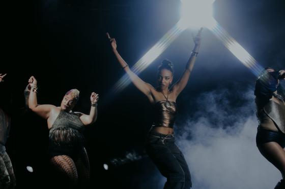 Mehrere Performer*innen tanzen vor schwarzem Hintergrund und weissem Bühnennebel unter grellem Gegenlicht. Im Zentrum hebt eine Person in einem dunklen, glänzenden Oberteil beide Arme hoch und blickt direkt nach vorne. Links und rechts sind weitere Körper und erhobene Arme teilweise angeschnitten zu sehen. Die Stimmung ist kraftvoll, clubartig und von starkem Lichtkontrast geprägt.