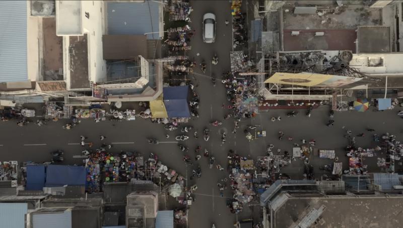 High aerial view of a street intersection filled with market activity. Stalls, tarps, and displays line both sides; many small figures move between the booths, and a car sits near the center. The crossing’s geometry organizes the bustling scene like a map.