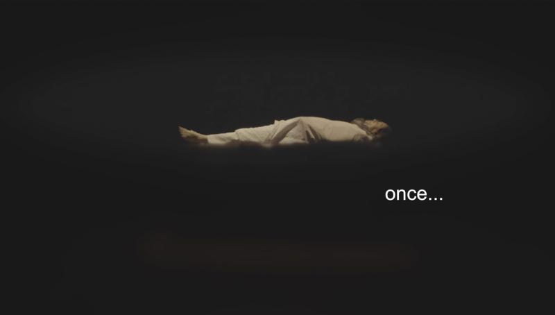 Dunkle, nebelige Bühne oder schwarzer Raum: In der Mitte liegt eine Person horizontal, fast wie schwebend im Schwarz. Rechts steht in weißer Schrift das Wort „once…“. Das Bild wirkt wie ein erzählerischer Einstieg, reduziert auf Körper, Raum und Text.