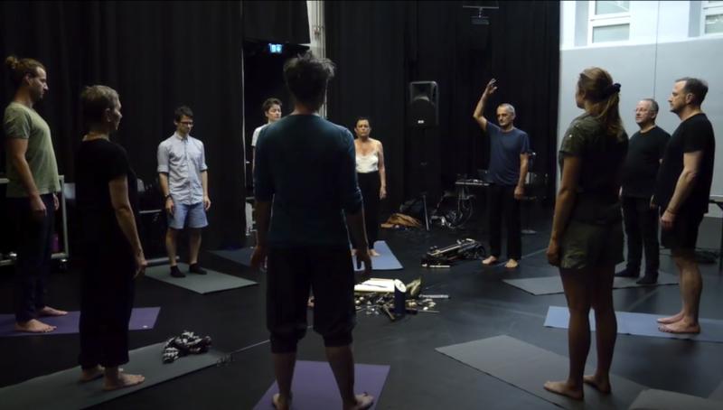 In einem dunklen, schwarzen Proberaum stehen mehrere erwachsene Personen barfuß auf einzelnen Yogamatten in einem offenen Kreis. Die Personen blicken zur Kreismitte, wo unterschiedliche Gegenstände wie Stöcke, Kabel und kleinere Objekte auf dem Boden liegen. Eine Person auf der rechten Seite hebt einen Arm nach oben, als würde sie eine Bewegung anleiten oder ein Zeichen geben. Der Raum wirkt wie ein Workshop- oder Probensetting für eine performative oder meditative Übung.