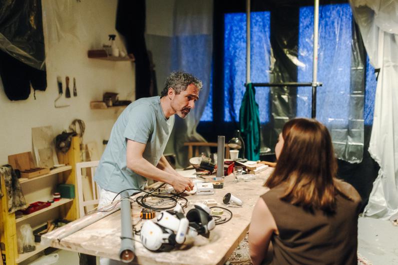 At a large worktable in a room that feels improvised and workshop-like, a person with curly grey hair leans over tools, cables and technical equipment. Opposite, a second person with their back to the camera appears seated or standing close by, listening. On the table are VR headsets, controllers, cables, tape and other components. In the background, dark fabrics and translucent plastic sheets hang in front of blue-lit windows.