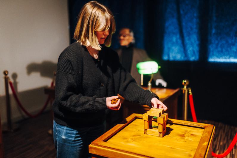 In the foreground, a person with a blonde bob and a dark knitted cardigan carefully builds a small structure from wooden blocks on a square wooden table. One hand holds another block while the other adjusts the top piece of the arrangement. In the background, slightly out of focus, a masked figure stands behind a desk with a green lamp, framed by red rope barriers. The light is warm and strongly concentrated on the table and the hands.