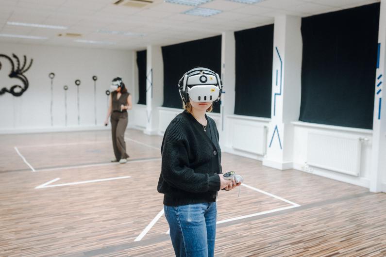 In einem hellen Raum mit Holzfussboden und weissen Wänden steht im Vordergrund eine Person mit VR-Headset und grossen Kopfhörern. Die Person trägt eine dunkle Strickjacke und Jeans und hält zwei Controller in den Händen. Weiter hinten bewegt sich eine zweite Person mit ähnlicher Ausrüstung durch den Raum. Schwarze Vorhänge verdecken die Fenster, blaue Markierungen an den Wänden und Linien am Boden strukturieren die Spielfläche.