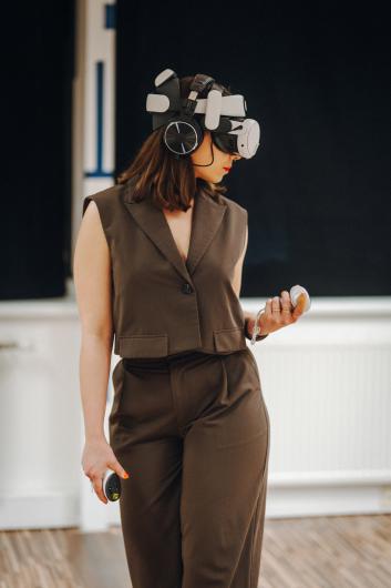 A person in a brown sleeveless trouser suit stands against a dark background wearing a large VR headset with headphones. They hold controllers in both hands, with their head angled downwards to the left behind the headset. The posture feels calm and concentrated, as if fully absorbed in a virtual environment. The pale floor and dark curtains make the equipment and the figure stand out clearly.