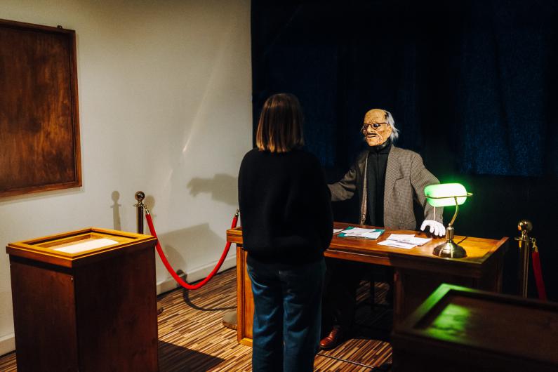 A person stands with their back to the camera in front of a small wooden table, facing a staged masked figure behind a desk. The figure wears a jacket, white gloves and an aged face mask; loose papers are scattered across the desk, and a small green lamp glows beside them. Red rope barriers mark off the area, giving it the feel of a museum display or official office. The scene suggests an encounter between a visitor and an uncanny bureaucratic installation.