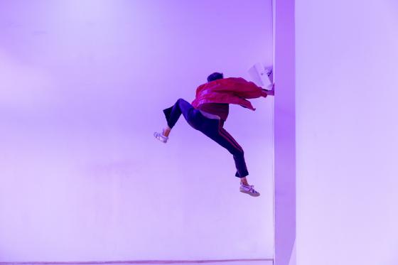 Eine Person in roter Jacke scheint seitlich an einer weißen Wand zu „klettern“, der Körper in einer dynamischen, schwebenden Pose, violett gefärbtes Licht.