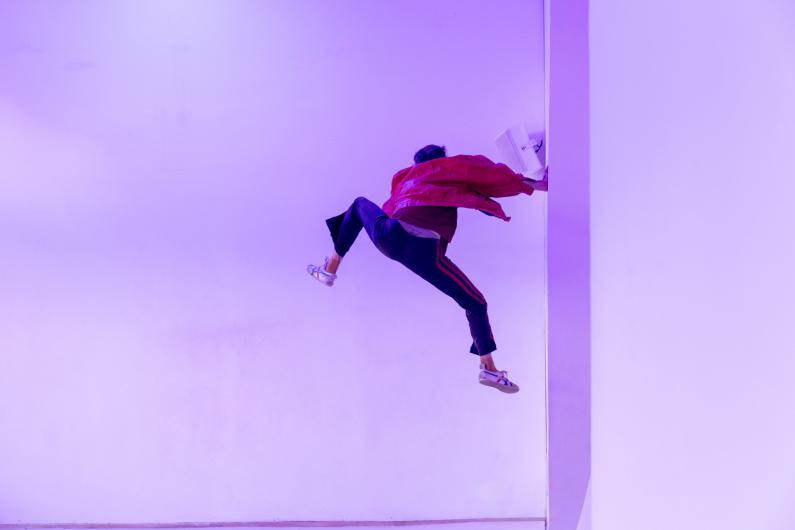 Eine Person in roter Jacke scheint seitlich an einer weißen Wand zu „klettern“, der Körper in einer dynamischen, schwebenden Pose, violett gefärbtes Licht.