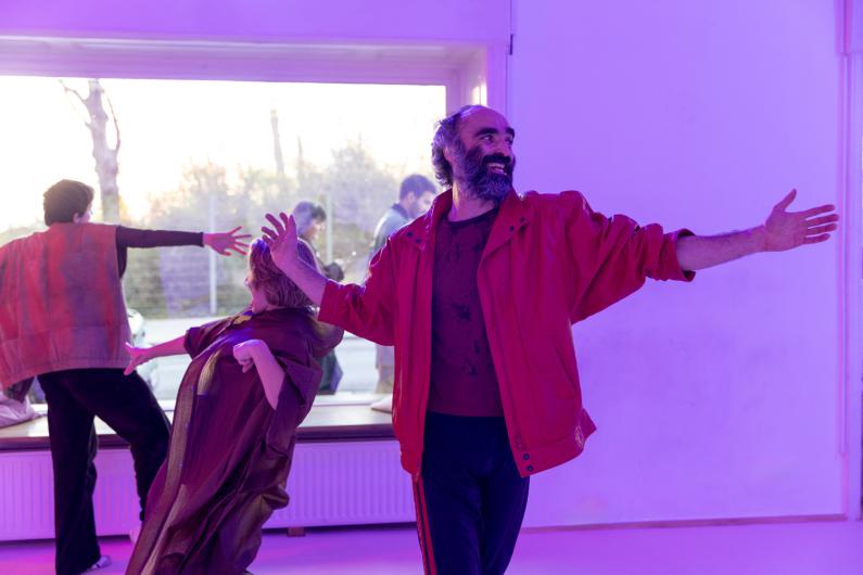 Several people move in a room in front of a large window; in the foreground, a person in a red jacket with arms outstretched, bathed in purple light.