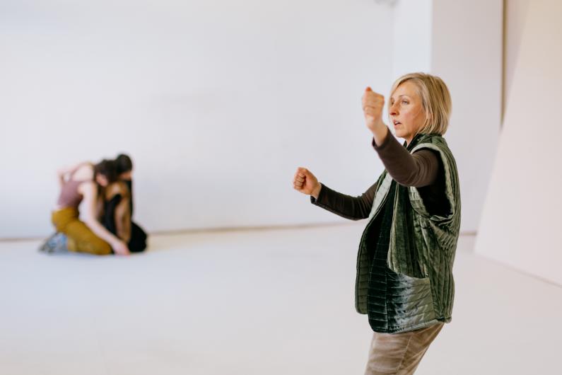 A person in a green quilted vest gestures with raised arms, while two blurred figures sit on the floor in the background of a bright space.